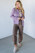 Woman wearing a purple cardigan, beige top, brown pants, and white shoes against a white brick wall.