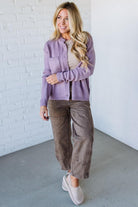 Woman wearing a purple jacket and brown pants against a white brick wall.