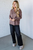 Woman wearing a brown cardigan and black pants against a white brick wall.