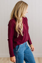 BLONDE WOMAN IN A BRIGHTLY LIT STUDIO WEARING DARK JEANS AND A SATIN RUCHED TRIM HALF BUTTON NECK, LONG SLEEVE, SOLID TOP IN BURGUNDY