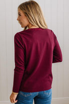 BLONDE WOMAN IN A BRIGHTLY LIT STUDIO WEARING DARK JEANS AND A SATIN RUCHED TRIM HALF BUTTON NECK, LONG SLEEVE, SOLID TOP IN BURGUNDY