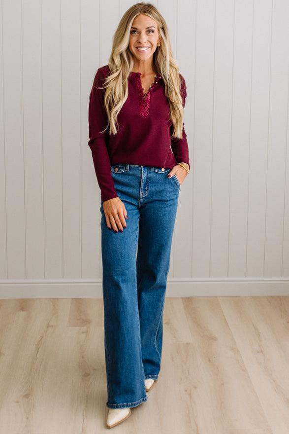 BLONDE WOMAN IN A BRIGHTLY LIT STUDIO WEARING DARK JEANS AND A SATIN RUCHED TRIM HALF BUTTON NECK, LONG SLEEVE, SOLID TOP IN BURGUNDY