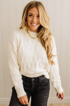 Woman wearing a white cable knit sweater and black jeans against a neutral background