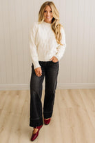 Woman wearing a white sweater, black jeans, and red shoes standing against a light wooden floor and wall.