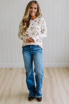 A woman stands in a bright room wearing a cream long-sleeve crewneck sweater with a brown floral pattern, blue jeans, and brown shoes.
