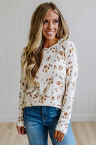 A woman with long blonde hair wearing a cream-colored knit sweater with a brown and tan floral pattern, paired with blue jeans, standing against a white paneled wall.