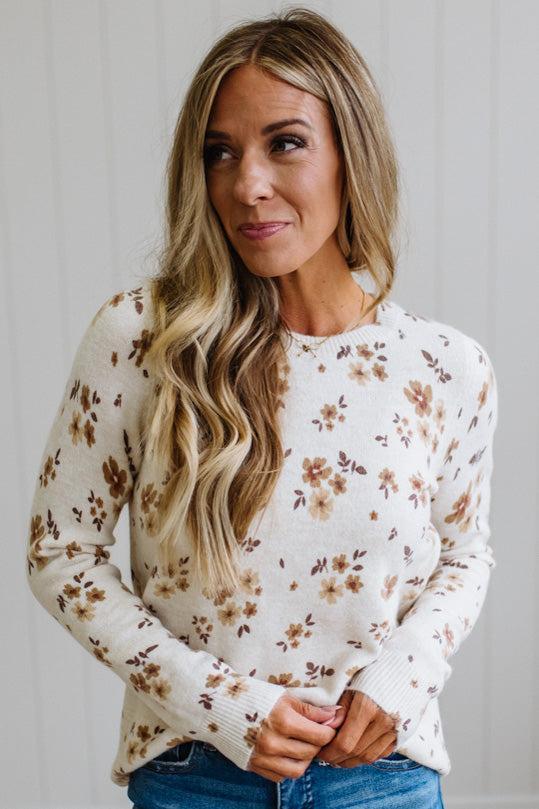 A woman with long blonde hair wearing a cream-colored knit sweater with a brown and beige floral pattern, paired with blue jeans, standing against a light gray paneled wall.