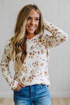 A woman with long blonde hair smiling while wearing a cream-colored long-sleeve knit sweater with a brown floral pattern, paired with blue jeans, standing against a white paneled wall.