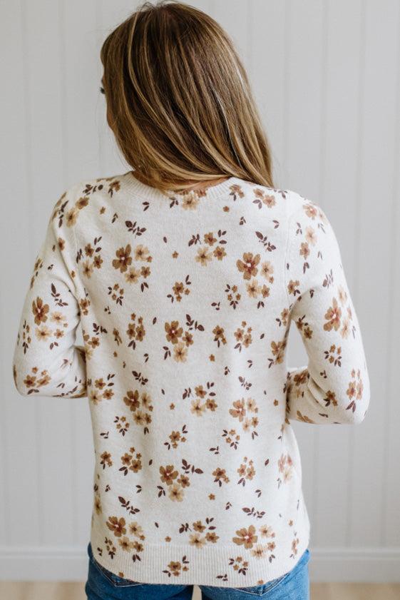 A woman with long blonde hair wearing a cream-colored knit sweater with a brown and tan floral pattern, paired with blue jeans, standing against a white paneled wall.