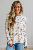 A woman wearing a cream-colored knit sweater with a brown and beige floral pattern, paired with blue jeans, standing against a white paneled wall.