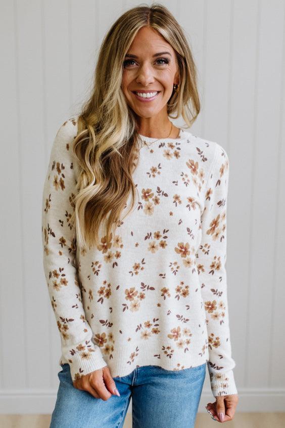 A woman wearing a cream-colored knit sweater with a brown and beige floral pattern, paired with blue jeans, standing against a white paneled wall.