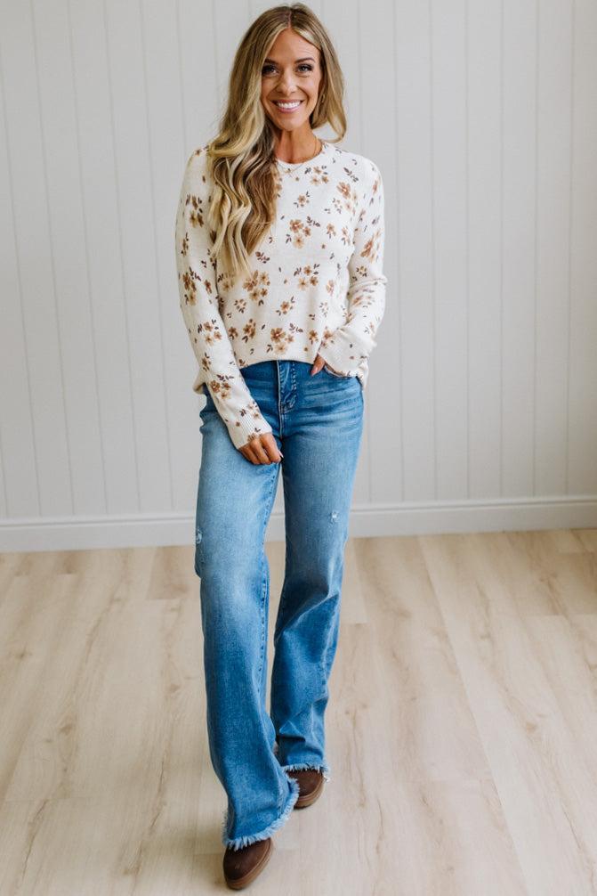 A woman stands in a bright room with white paneled walls and light wood flooring, wearing a cream long-sleeve crewneck sweater with a brown floral pattern, blue flare jeans, and brown boots.
