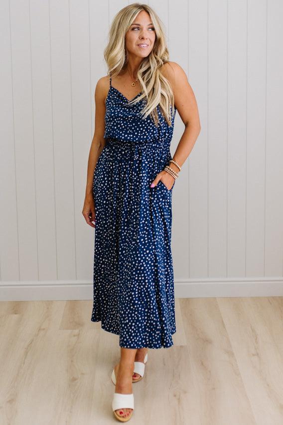 Woman wearing a navy blue dress with white polka dots against a light wooden floor and white wall background.