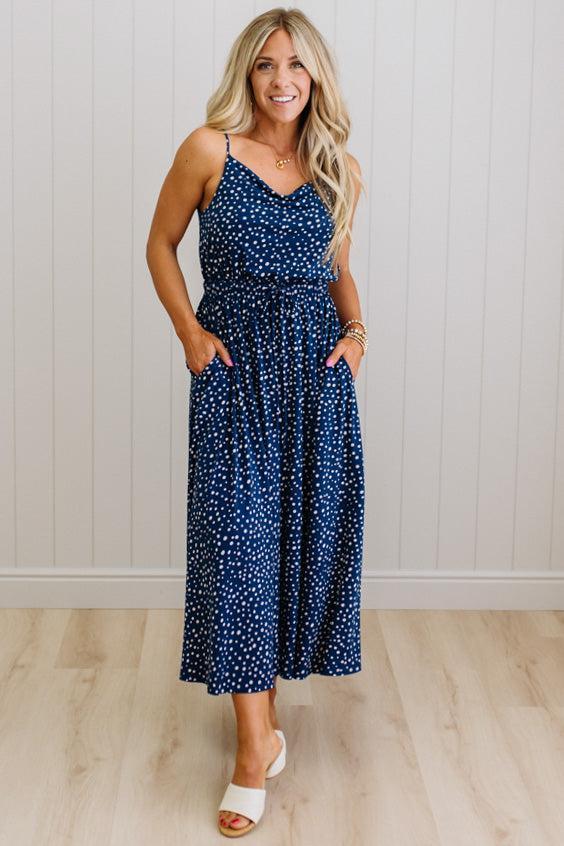 Woman wearing a navy blue dress with white polka dots against a light wooden floor and wall background.
