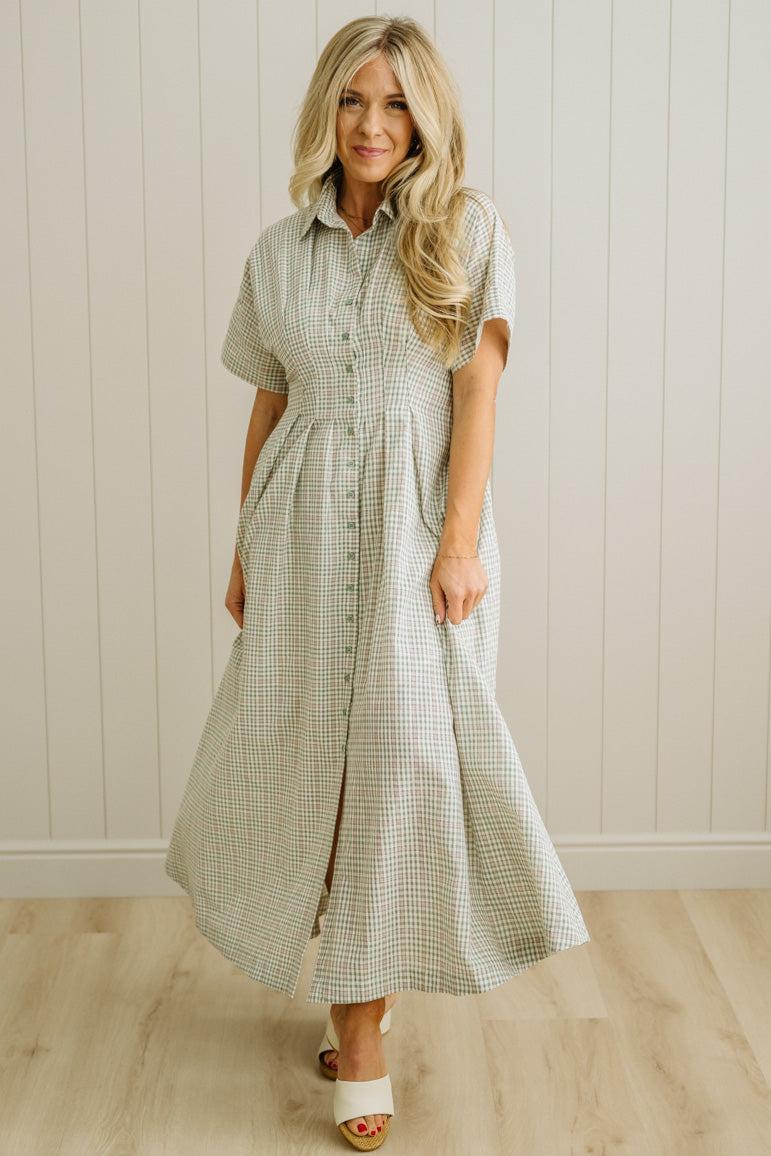 Woman wearing a green checkered dress standing against a white paneled wall.