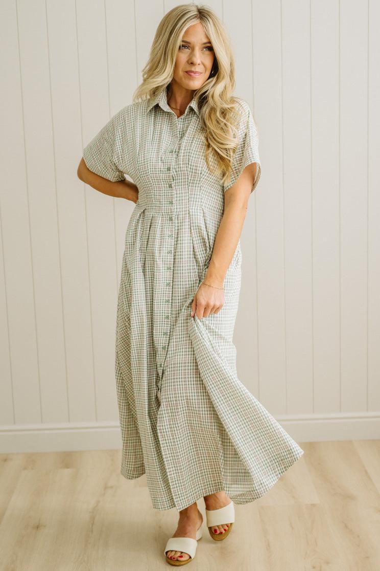 Woman wearing a green checkered dress standing against a white paneled wall.
