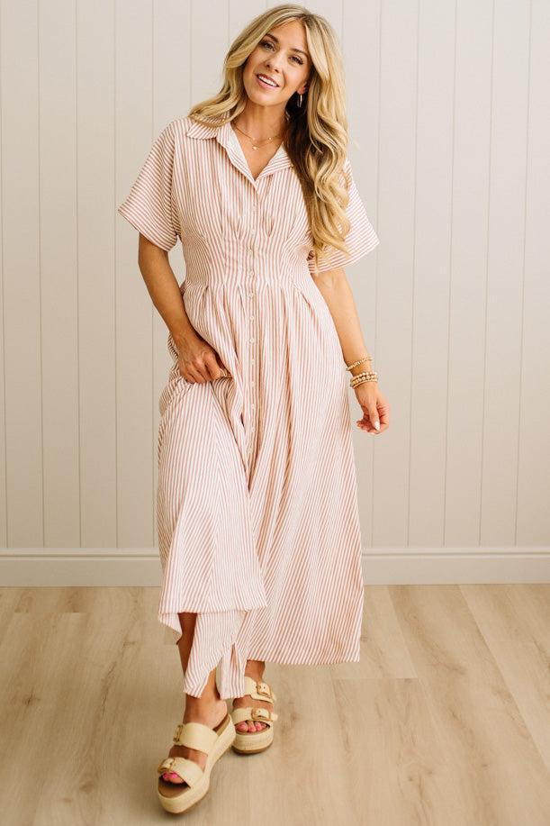 Woman wearing a pink and white striped dress standing against a light wooden paneled wall.
