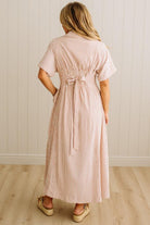 Woman wearing a long pink and white striped dress with a belt, standing against a light wooden paneled wall.