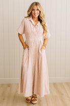 Woman wearing a long, light-colored striped dress against a white paneled wall.