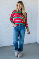 Woman wearing a red and green striped sweater with blue jeans against a white brick wall.