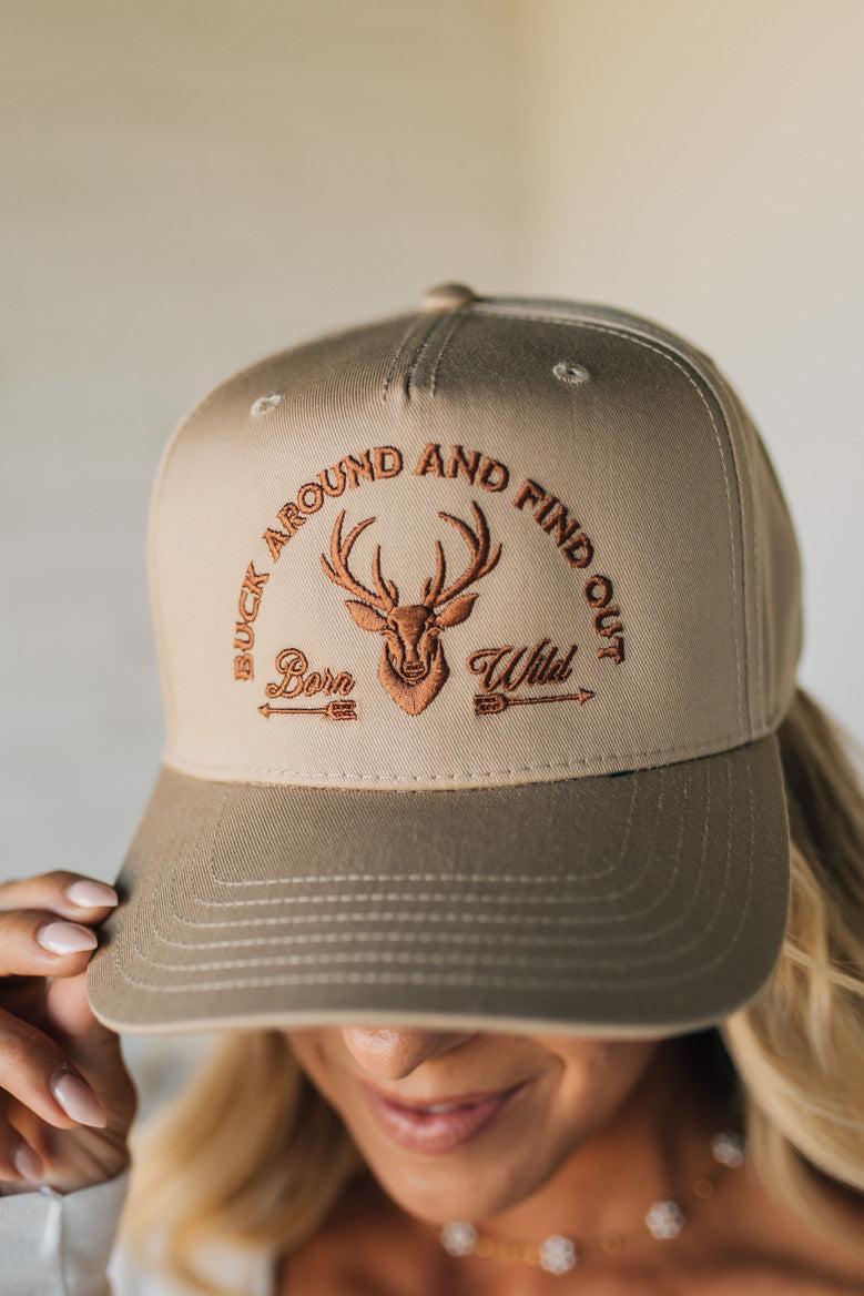 Beige cap with embroidered text and deer design held by a person against a neutral background
