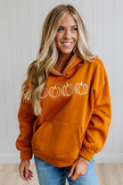 A smiling blonde woman wearing a burnt orange pullover hoodie with a white pumpkin graphic across the chest, paired with blue jeans, standing against a white paneled wall.