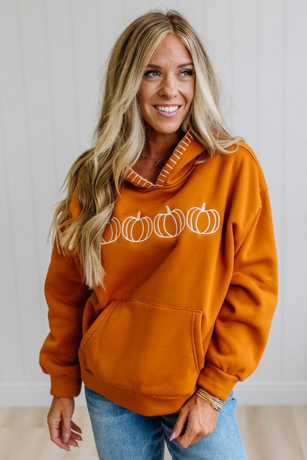 A smiling blonde woman wearing a burnt orange pullover hoodie with a white pumpkin graphic across the chest, paired with blue jeans, standing against a white paneled wall.