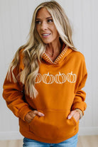 A woman wearing a burnt orange pullover hoodie with a white pumpkin graphic across the chest, paired with blue jeans, standing against a white paneled wall.