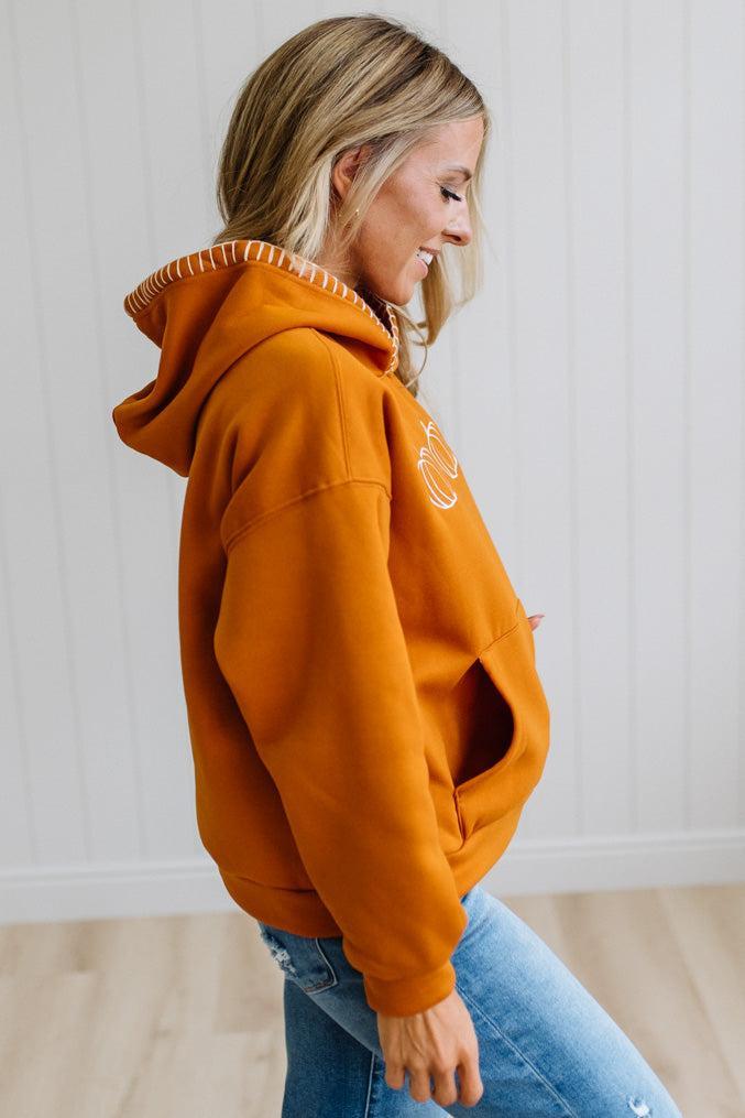 A woman wearing an oversized burnt orange hoodie with white embroidered details and a white striped trim along the hood, paired with blue jeans, standing against a white paneled wall.