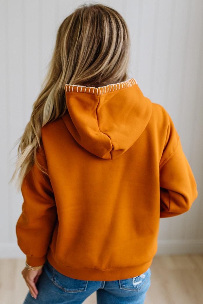 A woman viewed from behind wearing a burnt orange pullover hoodie with white contrast stitching along the hood's edge, paired with blue jeans.