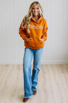 A woman wearing an oversized burnt orange hoodie with white embroidered details and a white striped trim along the hood, paired with blue jeans, standing against a white paneled wall.