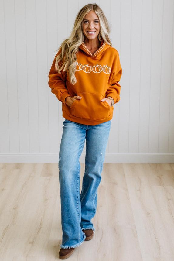 A woman wearing an oversized burnt orange hoodie with white embroidered details and a white striped trim along the hood, paired with blue jeans, standing against a white paneled wall.