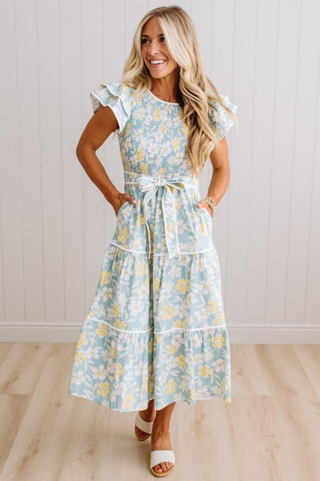 A woman modeling a light blue and yellow floral tiered maxi dress with ruffled cap sleeves and a matching waist tie, standing on a light wood floor against a white paneled wall.