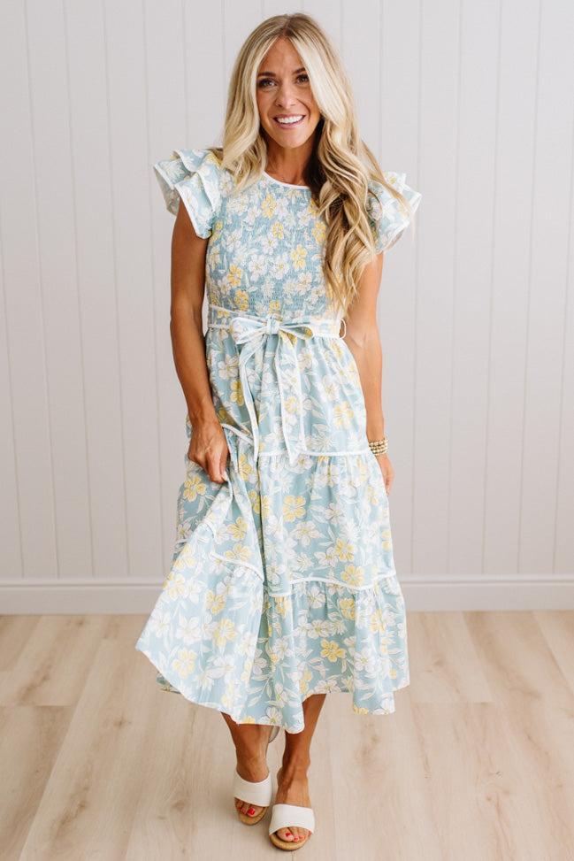 A woman modeling a light blue and yellow floral tiered midi dress with ruffled cap sleeves and a matching waist tie, standing on a light wood floor against a white paneled wall.