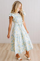 A woman modeling a light blue tiered midi dress with a yellow and white floral print, ruffled cap sleeves, and a tie waist, paired with white slide sandals in a bright room with light wood floors and white paneled walls.