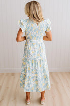 A woman viewed from behind wearing a light blue and yellow floral tiered midi dress with white ruffle sleeves, standing on a light wood floor against a white paneled wall.