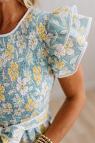 Close-up of a woman wearing a light blue smocked dress with a yellow and white floral print, featuring ruffled short sleeves and a tie belt at the waist.