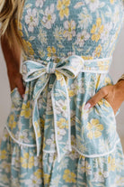 Close-up of a woman wearing a light blue floral dress with white and yellow flowers, featuring a matching fabric tie belt at the waist and side pockets.