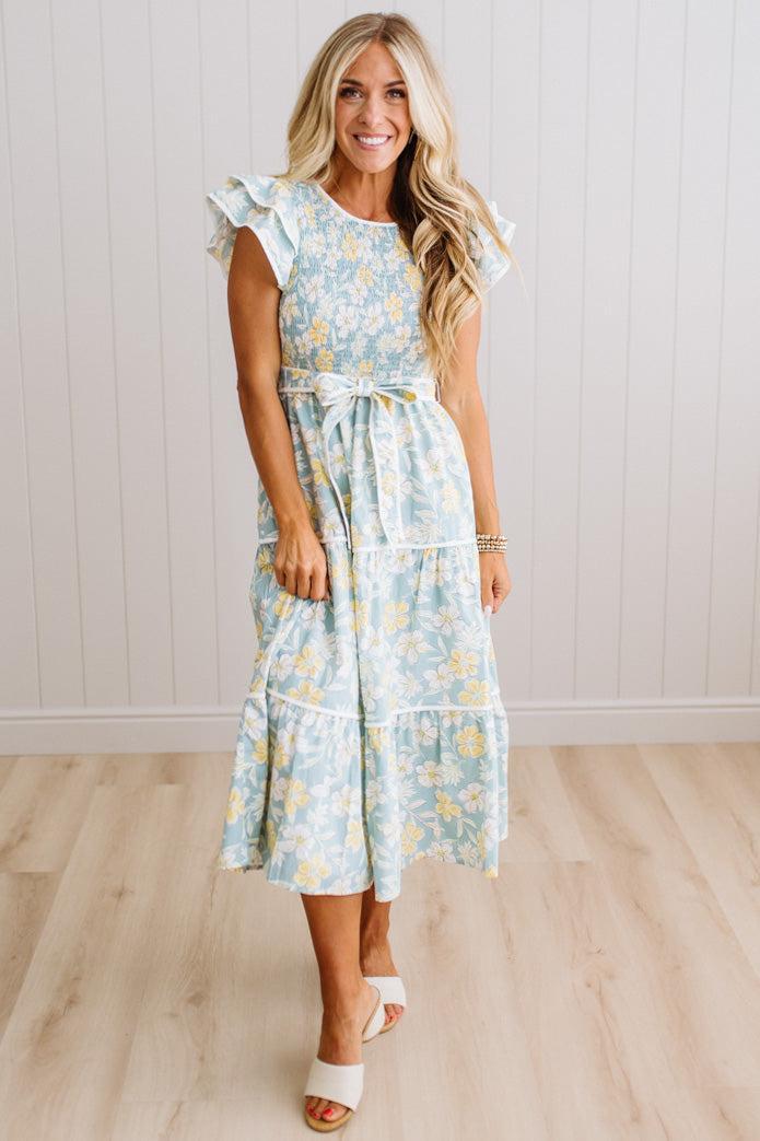 A woman modeling a light blue and white floral midi dress with ruffled cap sleeves and a matching fabric tie belt, standing in a bright room with light wood floors and white paneled walls.