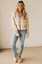 Woman wearing a floral cardigan, light blue jeans, and beige shoes against a white brick wall.