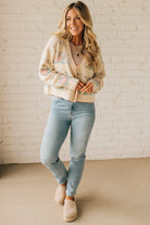 Woman wearing a patterned cardigan, light blue jeans, and beige shoes against a white brick wall.