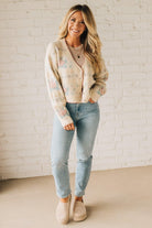Woman wearing a floral cardigan, light blue jeans, and beige boots against a white brick wall.