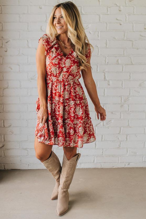 Woman wearing a red floral dress and beige boots against a white brick wall.