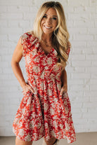 Woman wearing a red floral dress against a white brick wall.