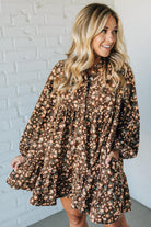 Woman wearing a brown floral dress against a white brick wall.