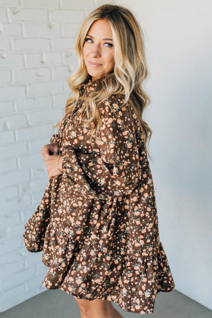 Woman wearing a brown floral dress against a white brick wall.