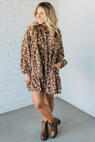 Woman wearing a brown floral dress with brown boots against a white brick wall.