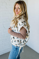 Woman wearing a floral-patterned vest over a long-sleeve shirt and jeans against a white brick wall.
