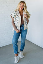 Woman wearing a floral vest, brown long-sleeve top, blue jeans, and white sneakers against a white brick wall.