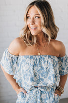 Blonde woman wearing a blue and white floral off the shoulder crop top with flowy overlay.